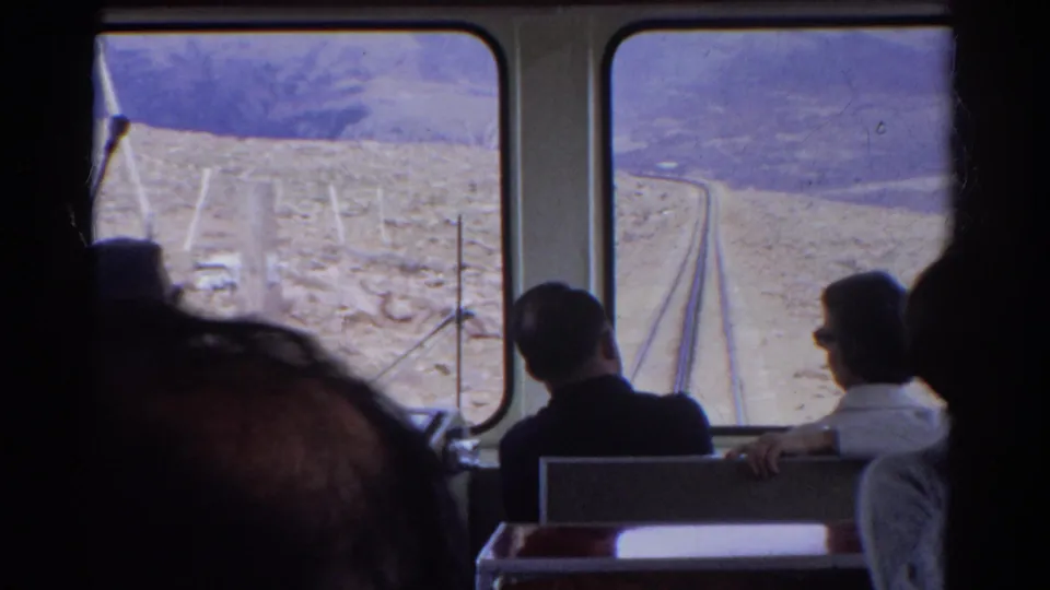 Passengers inside a 1967 Colorado train look out at tracks and arid mountain terrain in a grainy 8mm frame.