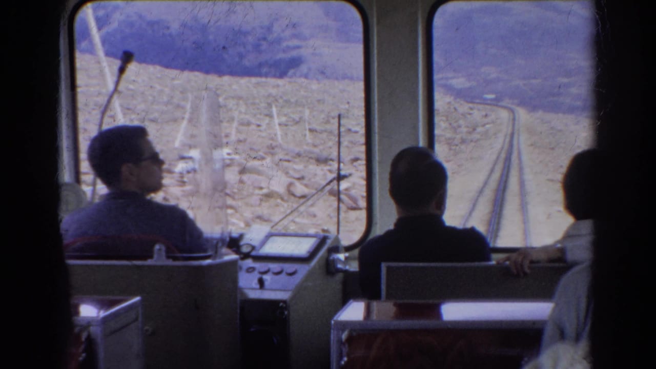 Man near the controls and other passengers face the front windows of a railway car.