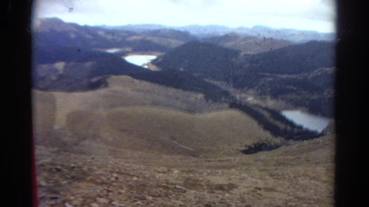 High overlook across a mountain valley with two pale lakes, rolling hills, forested ridges, and cloudy sky.