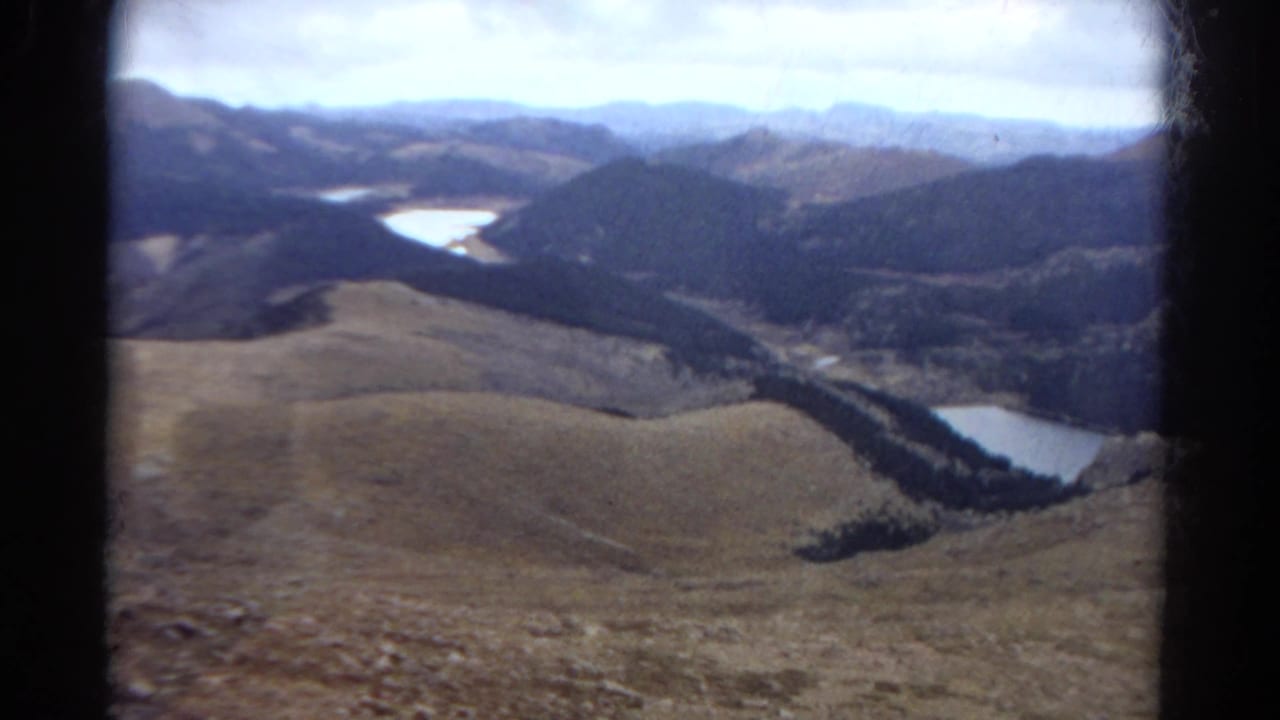 1967 Colorado home-movie frame showing a broad mountain valley, pale lakes, brown hills, and layered ridges within visible 8mm frame edges.