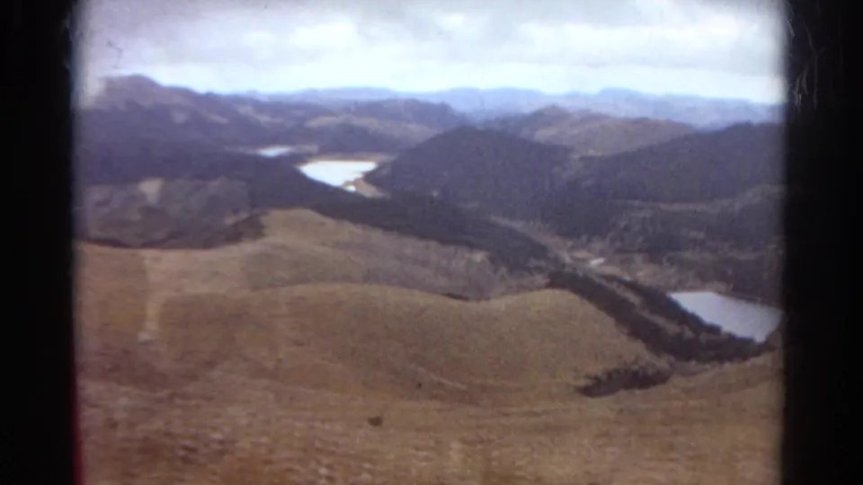 Colorado mountain landscape on 8mm film, with a sloped foreground hillside, a pale lake, wooded valley floor, and hazy mountain backdrop.