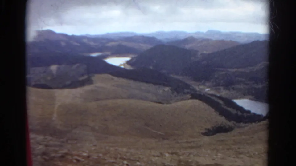 1967 Colorado 8mm frame of a wide mountain valley with lakes, rolling hills, dark forested ridges, and muted cloudy light.