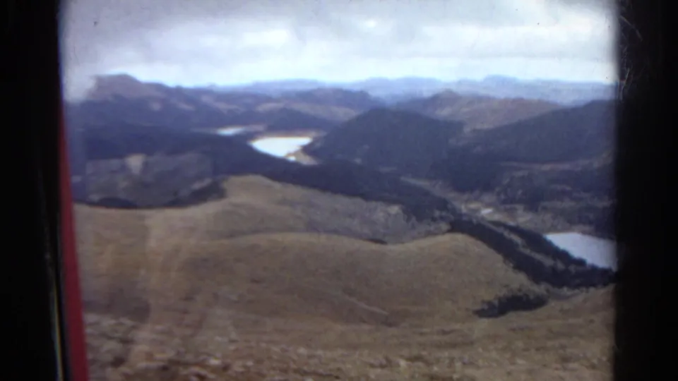 1967 Colorado 8mm view of rolling foothills, dark forested slopes, and pale water below, framed by a vehicle window.