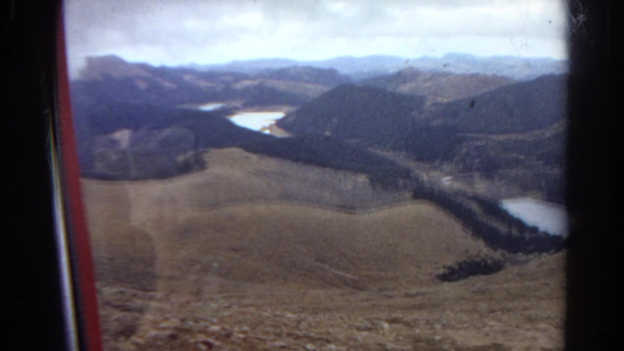 1967 Colorado home-movie frame showing a broad mountain valley with foothills, conifer-covered slopes, and bright water in the distance.