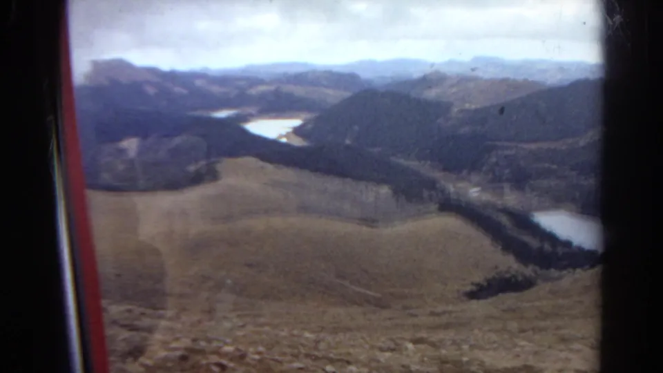 1967 Colorado home-movie frame showing a broad mountain valley with foothills, conifer-covered slopes, and bright water in the distance.