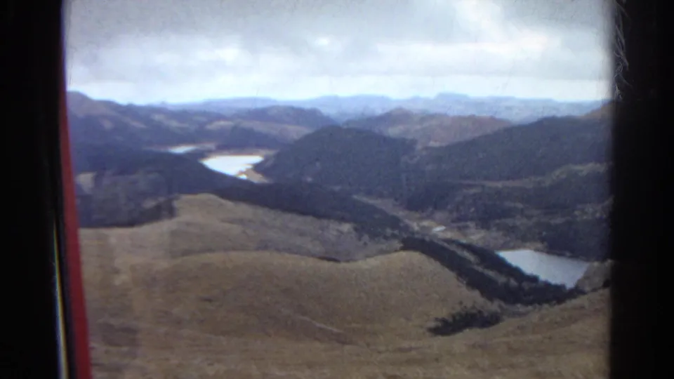 1967 Colorado 8mm footage frames a high view over brown foothills, wooded ridges, and a small lake or river bend.