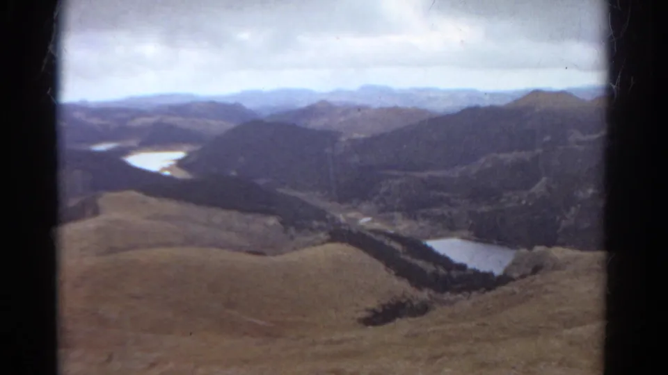 1967 Colorado travel footage shows rolling foothills, dark forested mountains, and water in a valley under muted daylight.