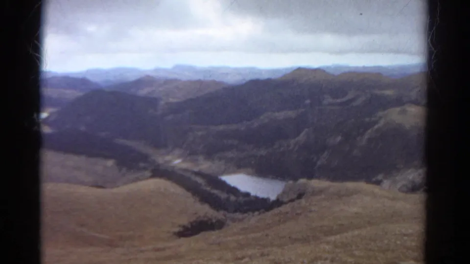 1967 Colorado home-movie frame showing a plane-window view across a mountain valley, forested ridges, and a bright lake.