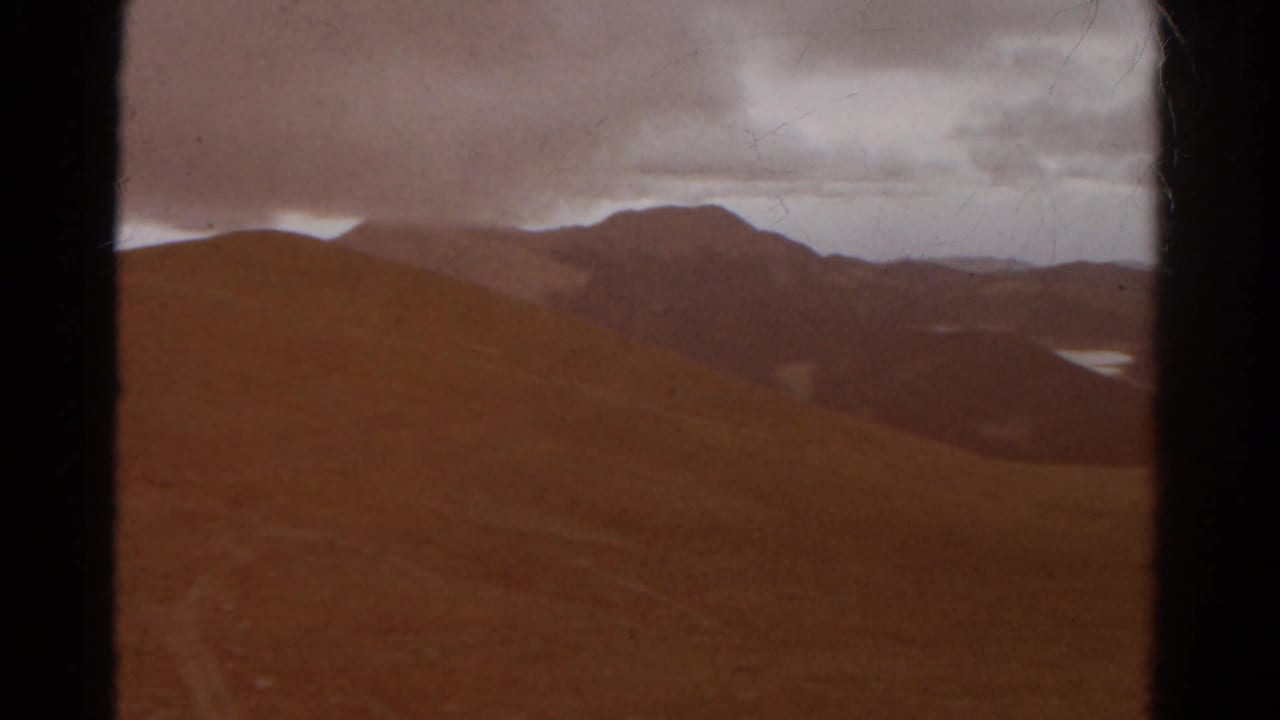 Colorado mountain ridges appear in soft 1967 8mm color, with a distant lake visible through the valley under a cloudy sky.