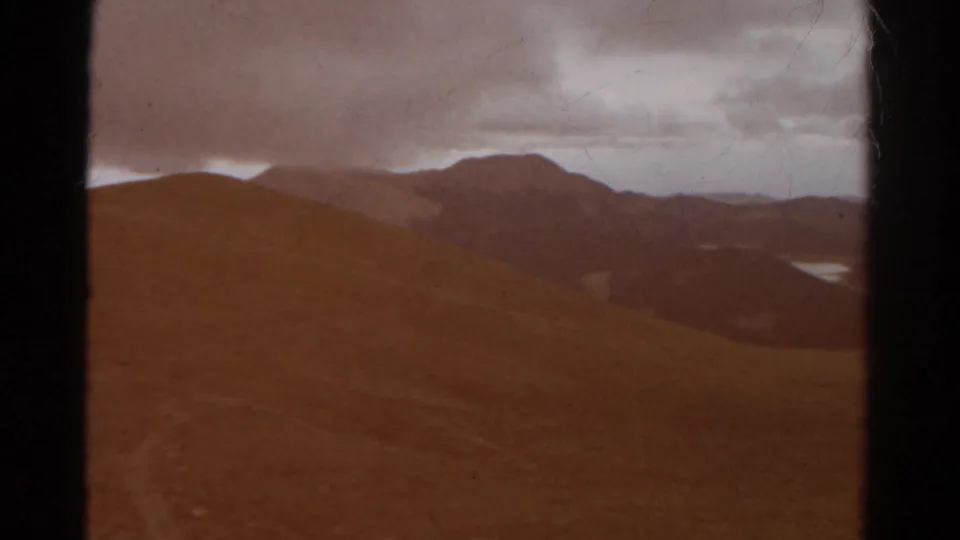Colorado mountain ridges appear in soft 1967 8mm color, with a distant lake visible through the valley under a cloudy sky.