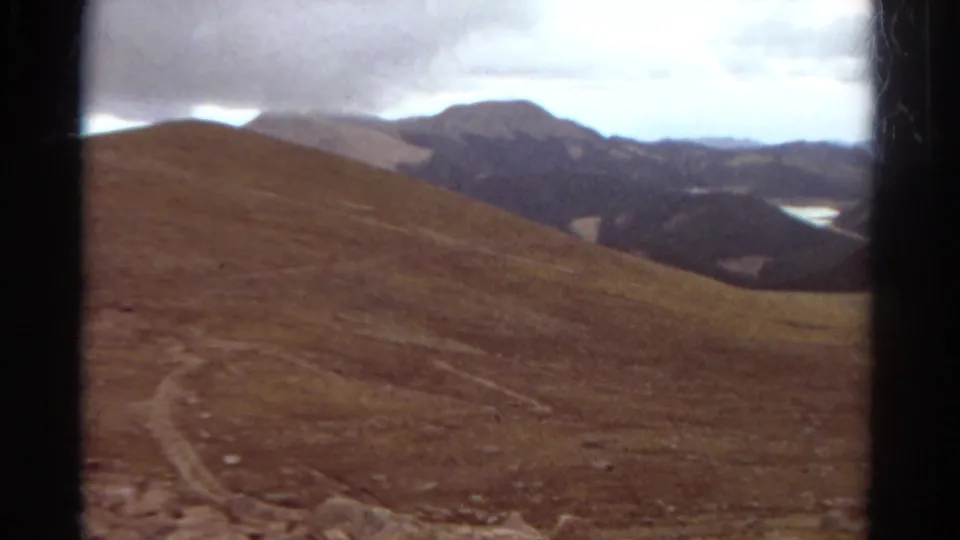 The 1967 Colorado home-movie frame shows a sloping hillside, distant mountains, and bright patches of lake water under cloud cover.