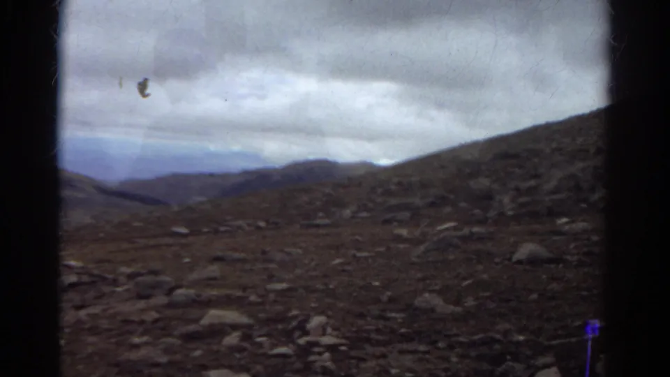 1967 Colorado 8mm view of scattered rocks, rough upland ground, a sloping hillside, and blue-gray ridges in the distance.