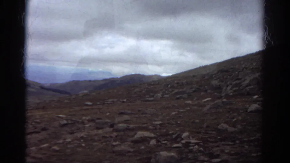 1967 Colorado home-movie still showing barren rocky ground leading toward a dim valley and layered ridges below heavy clouds.