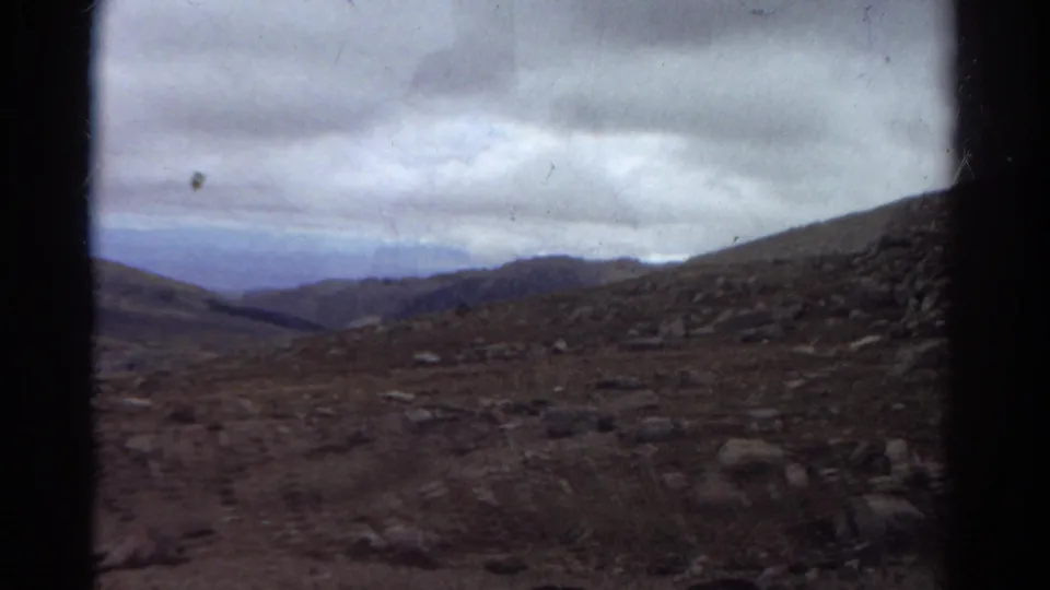 1967 Colorado 8mm frame with rough ground, scattered stones, a sloping hillside, distant mountains, and dark projection borders.