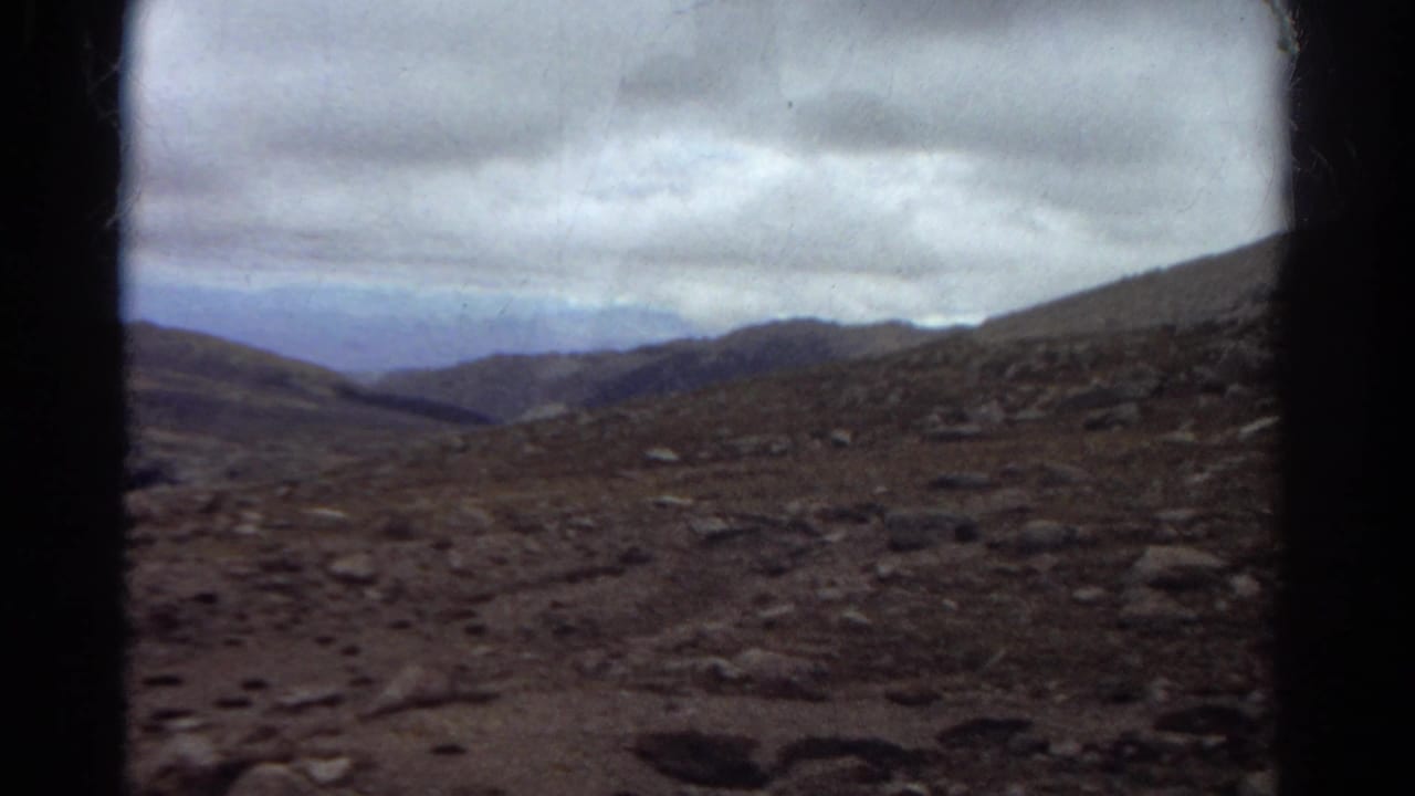 Rocky mountain slope with distant ridges under a cloudy sky, framed by dark 8mm film edges.