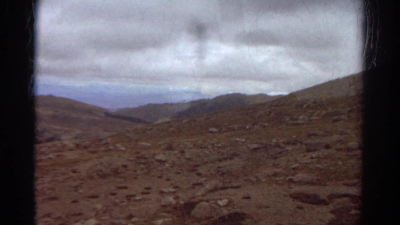 Brown rocky hillside and distant mountain valley beneath a gray, cloud-filled sky.