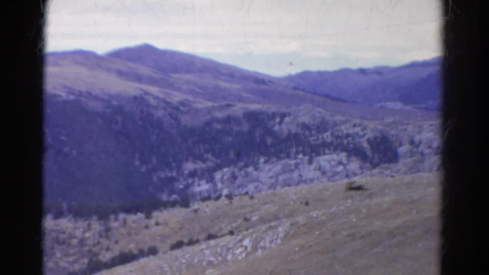 1967 Colorado 8mm frame showing blue-toned mountain ridges, sparse trees, rocky slopes, and visible film edges.