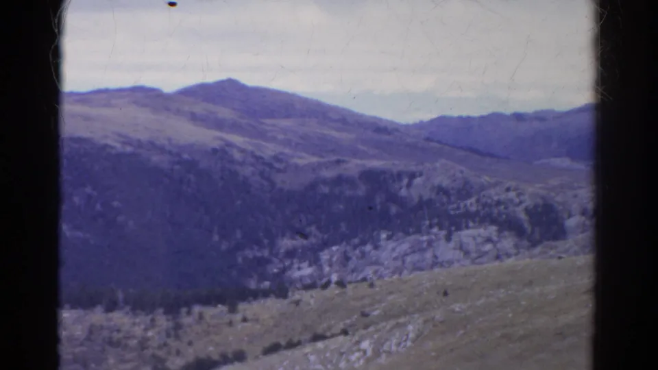 1967 Colorado 8mm view of rolling mountain ridges, evergreen-covered slopes, pale rock faces, and cloudy sky with visible film wear.