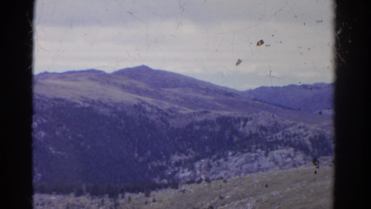 Colorado mountain landscape on 8mm film, showing layered ridges, tree-covered slopes, pale rocks, cloudy sky, and aged color shift.