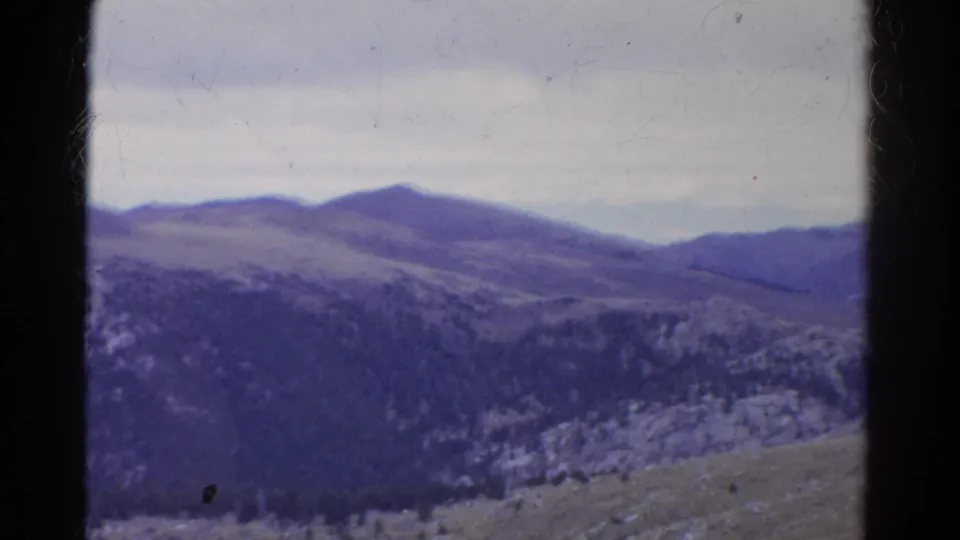 1967 Colorado home-movie frame of rolling mountain terrain, dark tree cover, rocky patches, and overcast sky with purple film aging.