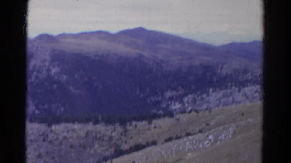Aged 8mm Colorado mountain view with dark evergreen slope, pale rock outcrops, open hillside, cloudy sky, and visible film wear.