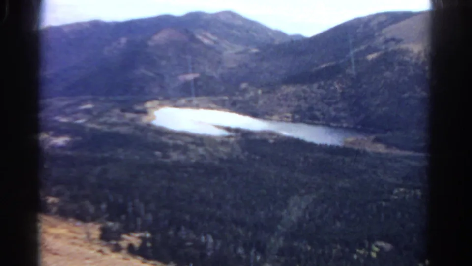 Wide 1967 Colorado home-movie frame showing a lake below evergreen slopes and rounded mountain peaks.