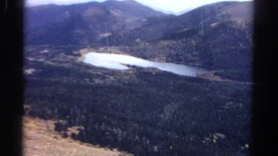 End frame of a 1967 Colorado 8mm scenic view, with a bright lake bordered by forest and mountain slopes.
