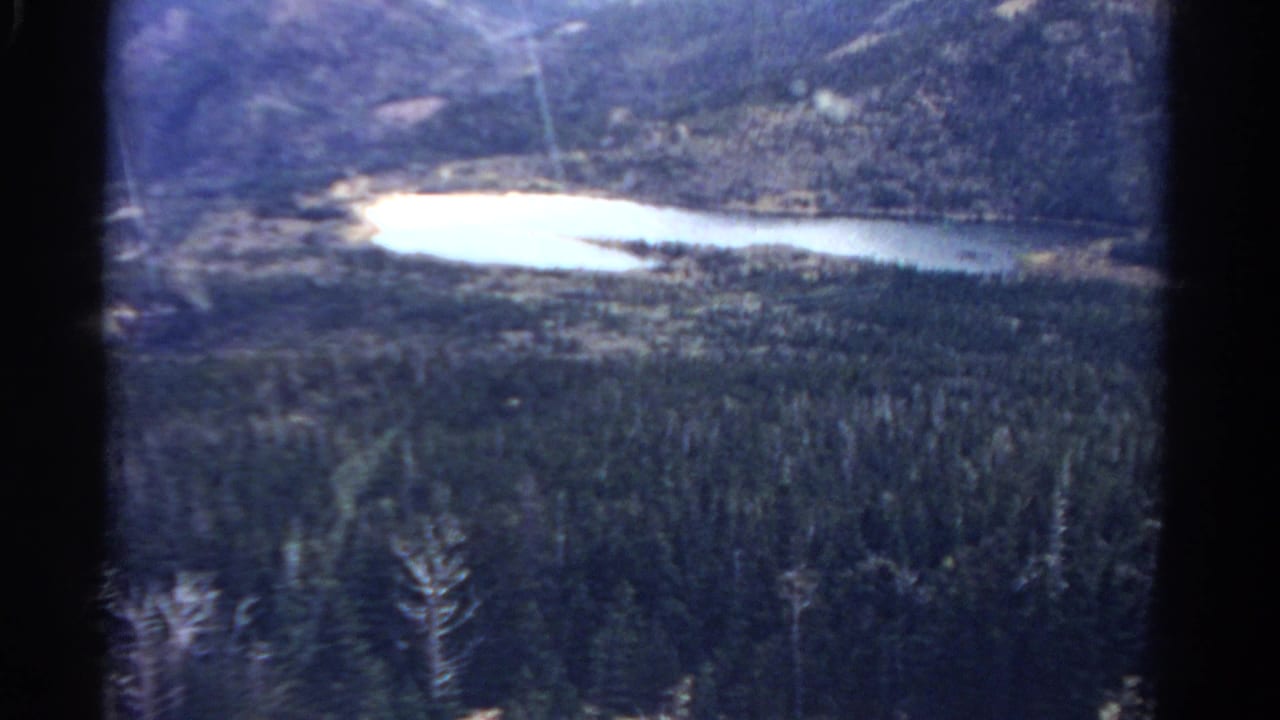 1967 Colorado 8mm frame showing a bright lake below forested mountain slopes from an elevated viewpoint.