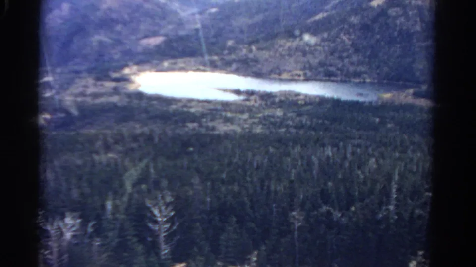 1967 Colorado 8mm frame showing a bright lake below forested mountain slopes from an elevated viewpoint.