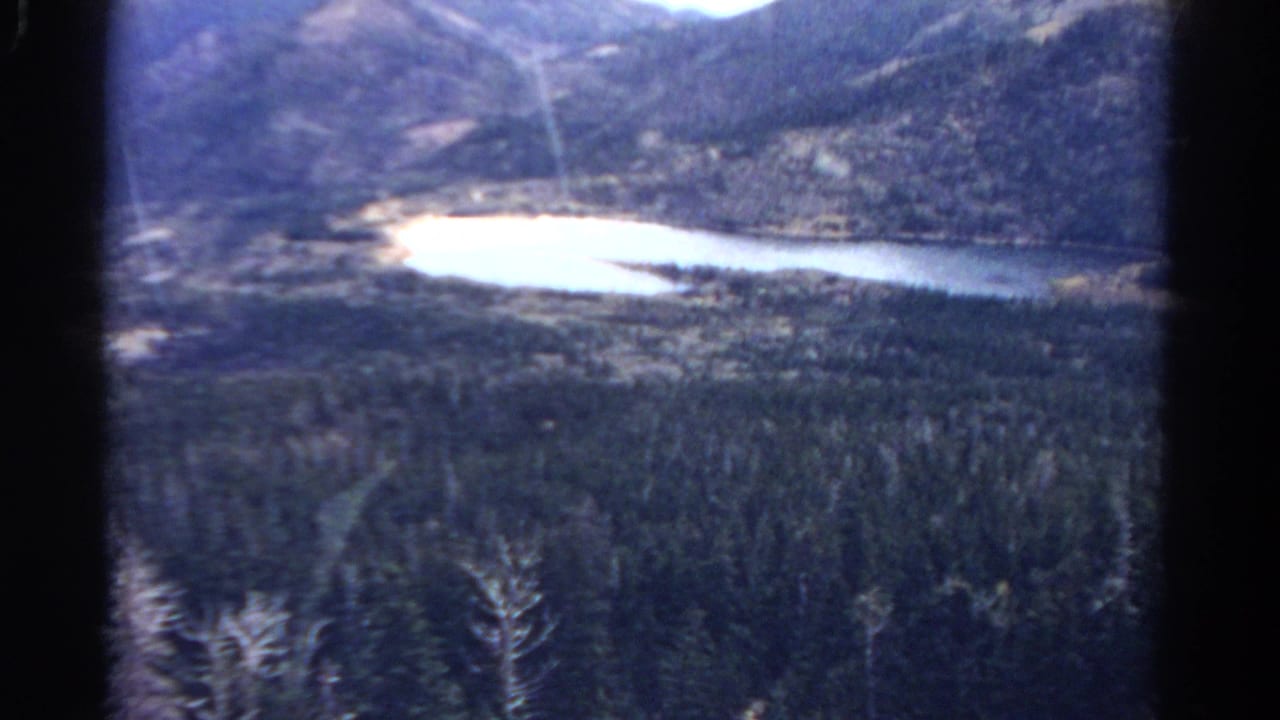1967 Colorado home-movie still with a reflective lake, wooded valley, and rugged mountain backdrop.