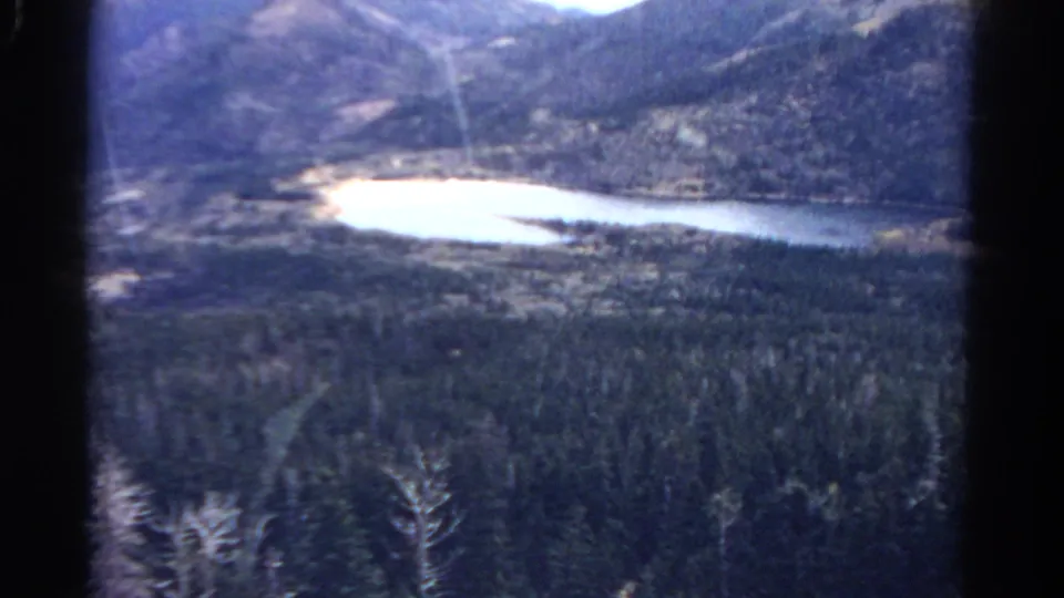 1967 Colorado home-movie still with a reflective lake, wooded valley, and rugged mountain backdrop.