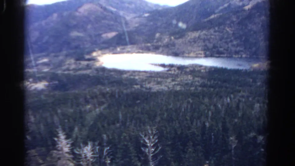 1967 Colorado 8mm frame with lake, evergreen forest, mountain terrain, and visible dark film-frame edges.