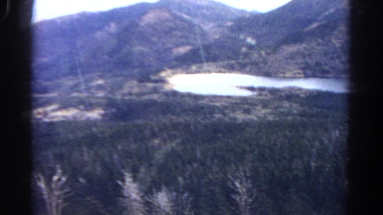 1967 Colorado 8mm frame showing a soft, elevated view of forested slopes, rounded mountains, pale sky, and a small lake below.