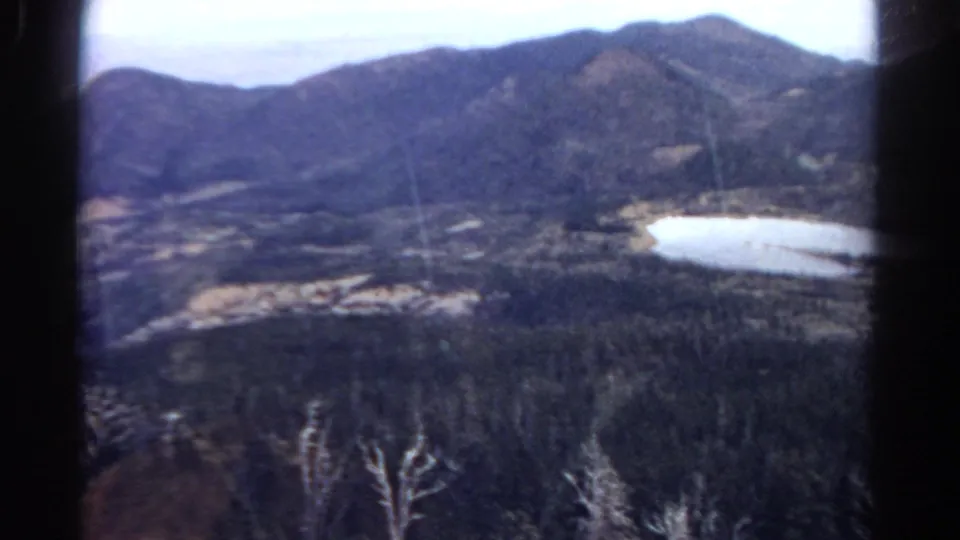 1967 Colorado 8mm frame showing a high view over wooded terrain toward a small lake and surrounding mountain slopes.
