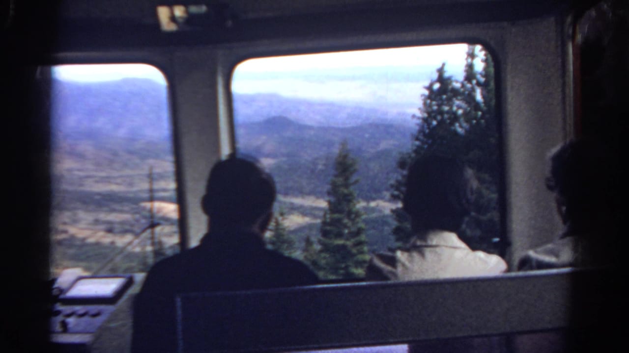 1967 Colorado 8mm frame of two silhouetted passengers seated in a tram cabin, looking out toward mountains, evergreens, and a valley.