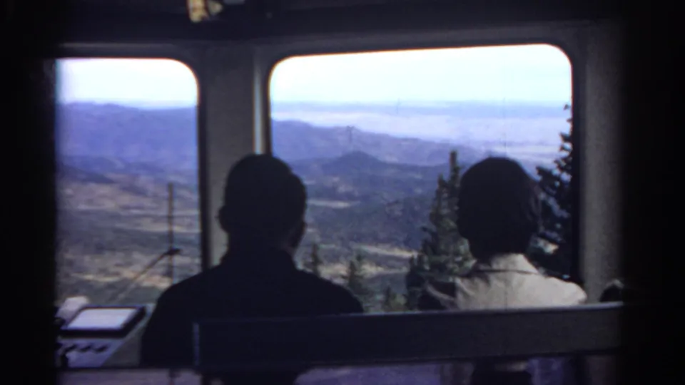 1967 home-movie frame from inside a Colorado tram, with seated riders in shadow and a broad mountain landscape beyond the windows.