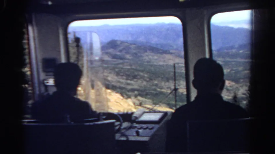 Inside the 1967 Colorado travel vehicle, two seated figures face a bright mountain slope and valley framed by windows.