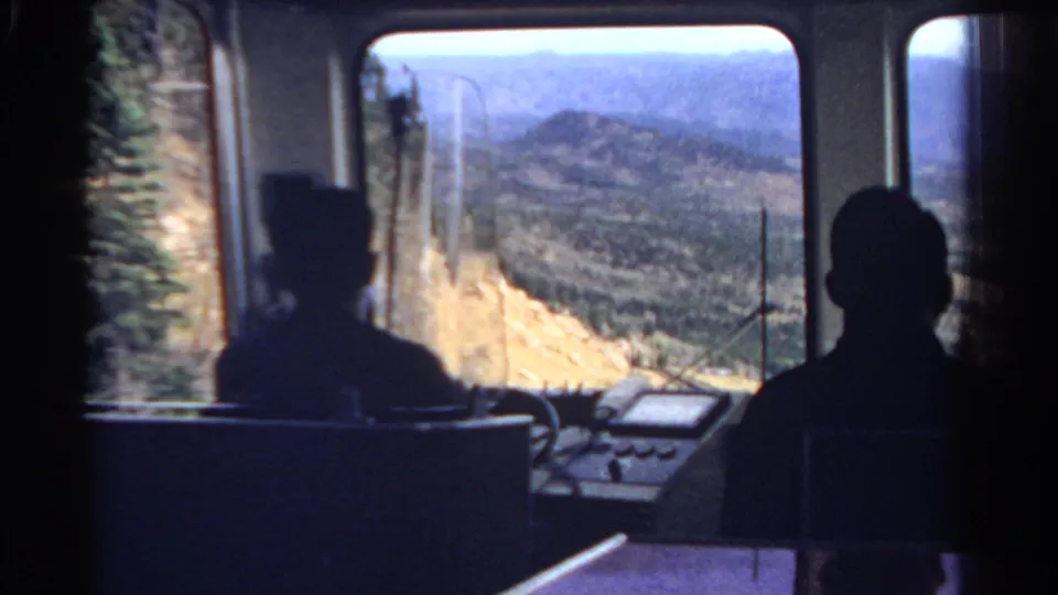 A seated passenger silhouette is framed against a broad Colorado mountain view from inside a windowed scenic vehicle.