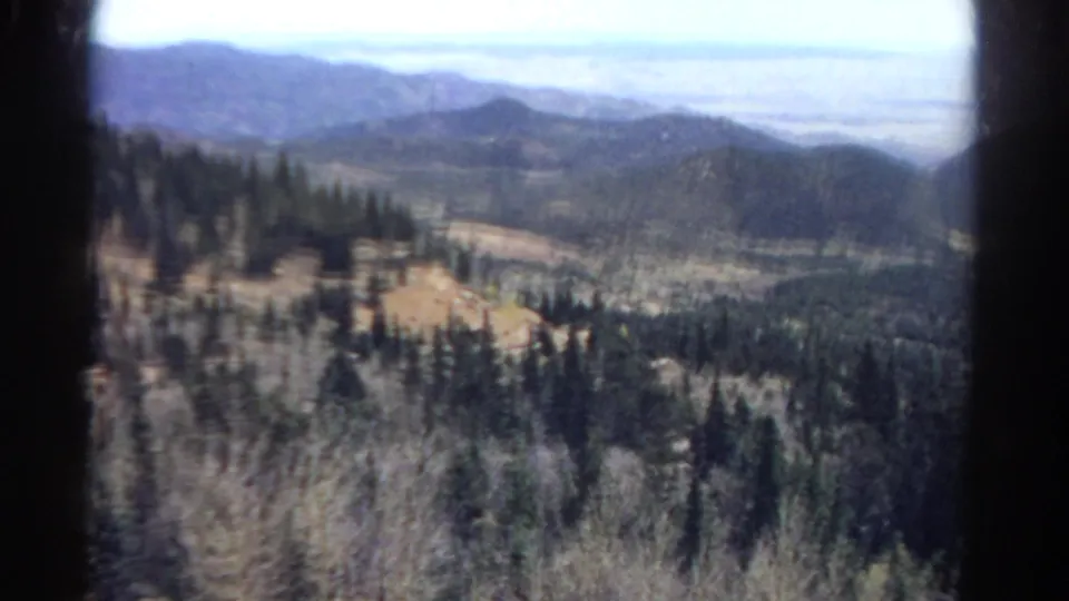 1967 Colorado mountain scenery through a train window, with pine-covered slopes and hazy distant ridgelines.