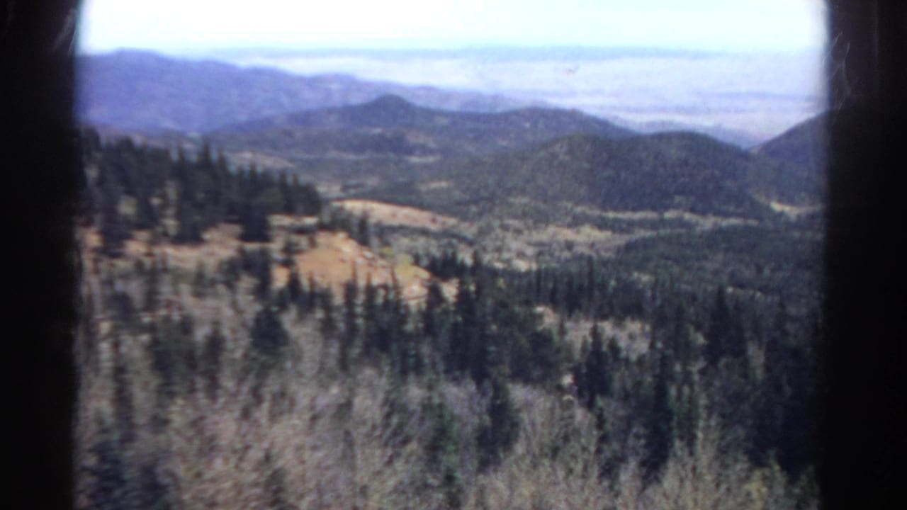 1967 Colorado 8mm frame showing a forested mountain valley, distant blue hills, dry foreground brush, and dark film-edge borders.