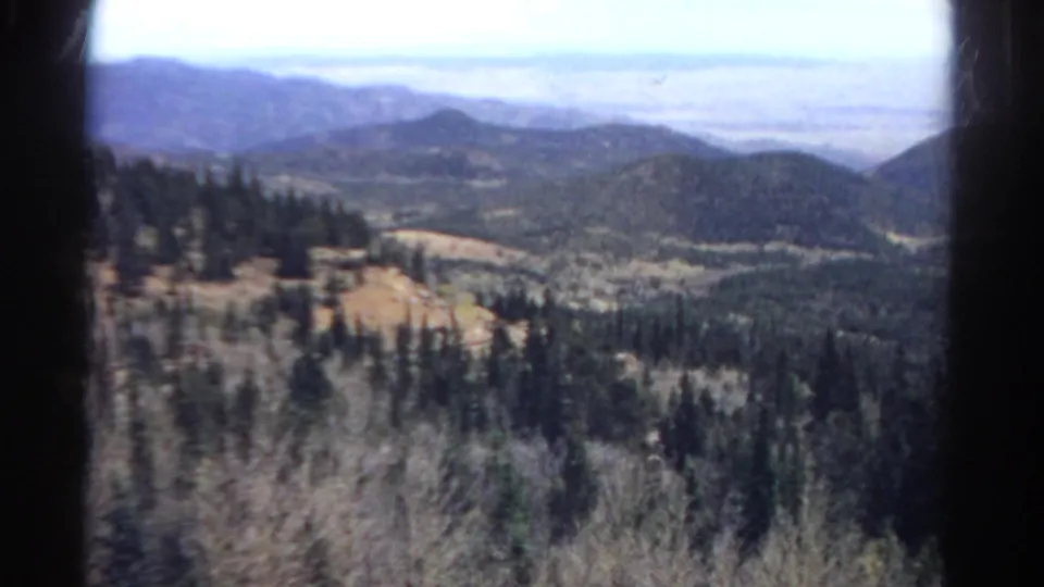 1967 Colorado 8mm frame showing a forested mountain valley, distant blue hills, dry foreground brush, and dark film-edge borders.