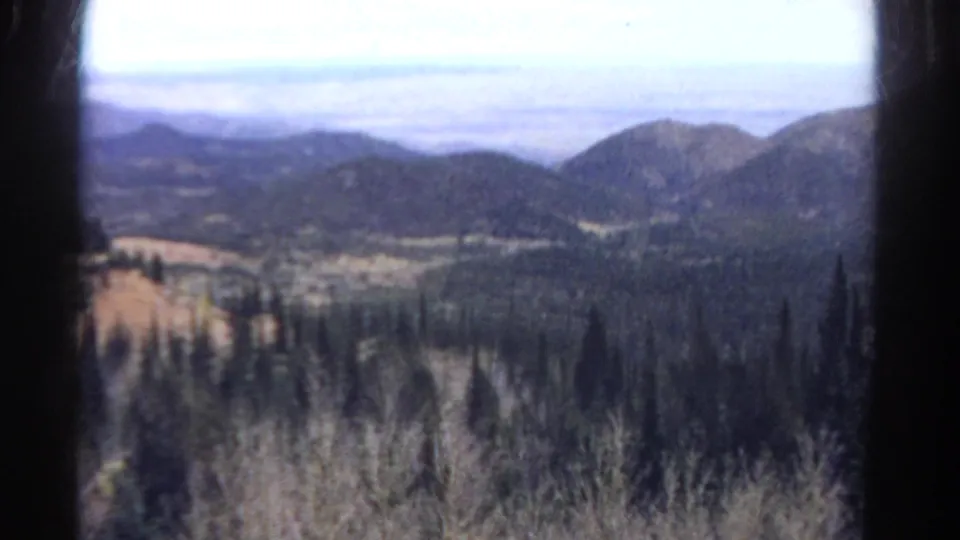 1967 Colorado landscape frame with soft-focus forest, foreground brush, rounded mountain slopes, and a hazy distant horizon.
