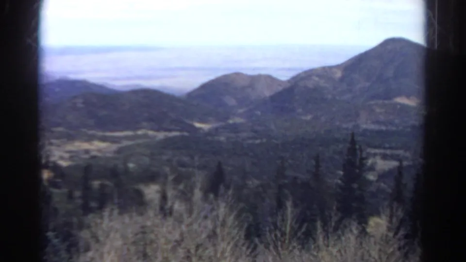 1967 Colorado 8mm frame looking over a forested valley toward rounded mountain ridges beneath a bright pale sky.