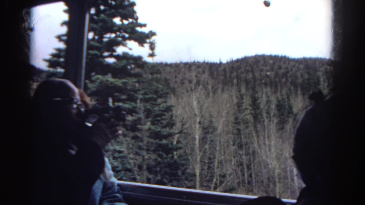 1967 Colorado 8mm frame showing tourists inside a train, with a man filming through the window toward evergreen trees and low mountain hills.