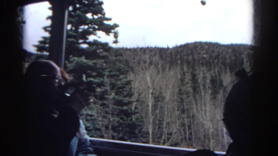 1967 Colorado 8mm frame showing tourists inside a train, with a man filming through the window toward evergreen trees and low mountain hills.