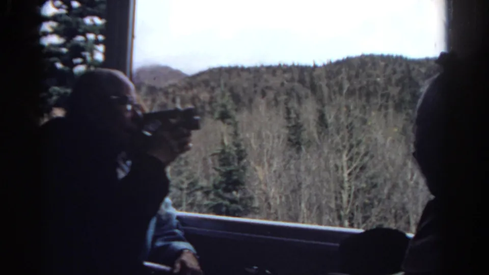 A shadowed passenger in a Colorado train carriage holds a camera near the window as wooded hills and evergreens move past outside.
