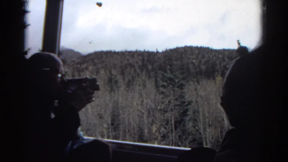 1967 Colorado 8mm frame showing a dark helicopter interior with a seated silhouette near the window and wooded mountain terrain outside.