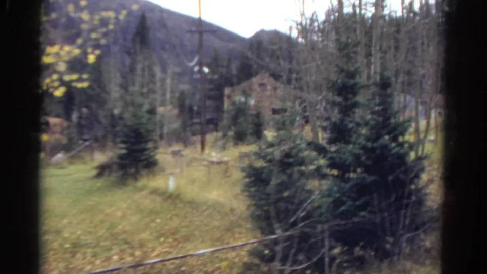 1967 Colorado 8mm frame showing a cabin-like building among trees, utility poles, brush, and mountain terrain seen from the moving helicopter.