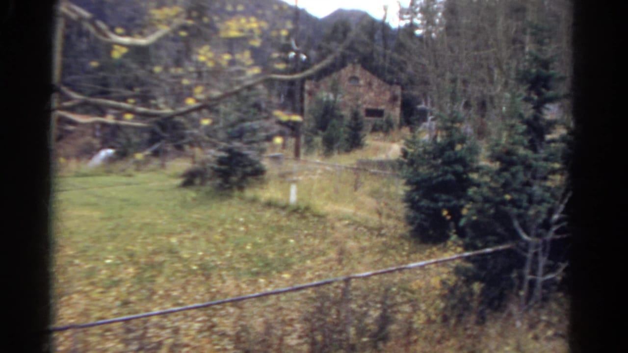 1967 Colorado 8mm frame showing a fenced rural yard with trees, a distant stone building, and mountains seen from behind a window edge.