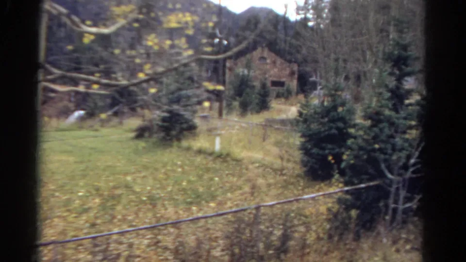 1967 Colorado 8mm frame showing a fenced rural yard with trees, a distant stone building, and mountains seen from behind a window edge.